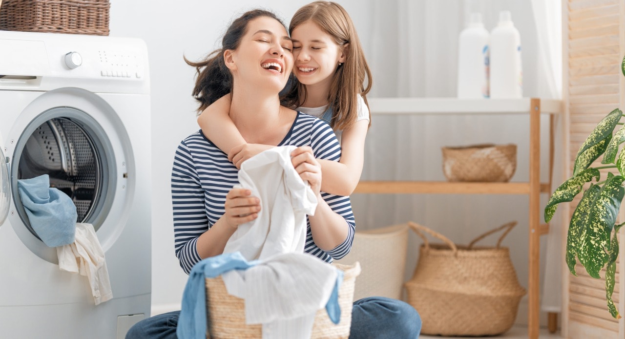 Mãe e filha recolhem roupas da máquina lavadas com sabão biodegradável, em um momento de cuidado e alegria na lavanderia.