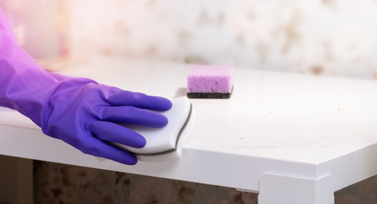 Pessoa com luvas roxas usando uma esponja branca para limpar uma superfície com desinfetante de lavanda