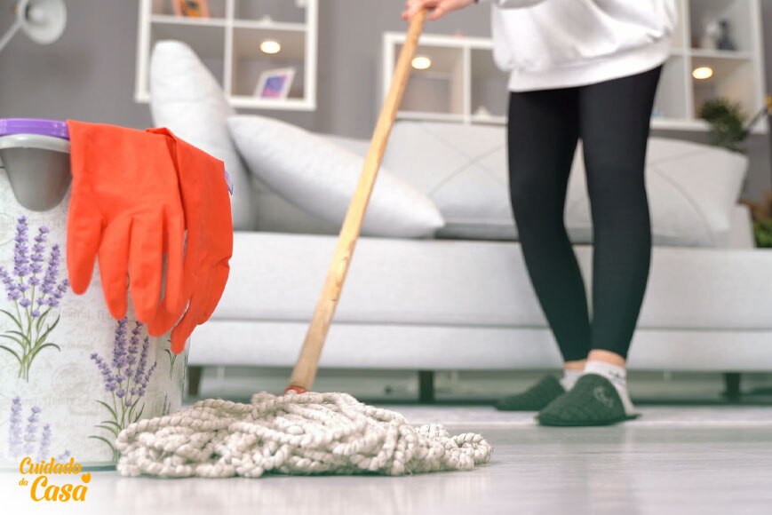 Aprenda a limpar a suMulher limpando a sala da casa, com um balde, luvas, rodo e produtos para limpar piso encardido.
