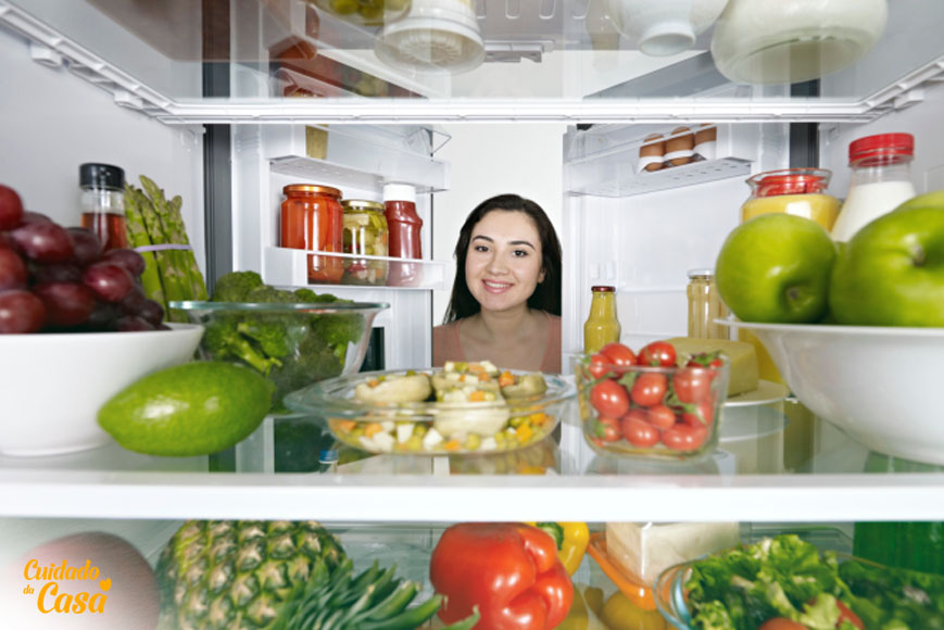 Mulher com a geladeira aberta olhando para vários alimentos e procurando como tirar mau cheiro da geladeira